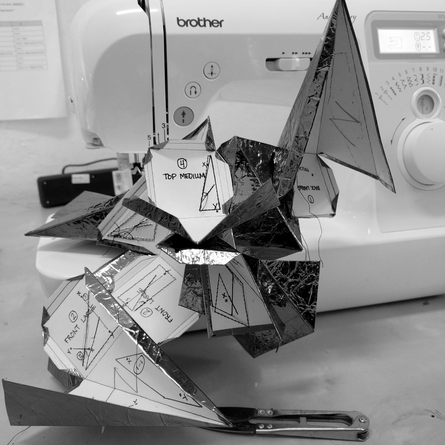 A metallic, geometric sculpture made of paper patterns sits near a sewing machine, showcasing intricate folding and design plans.