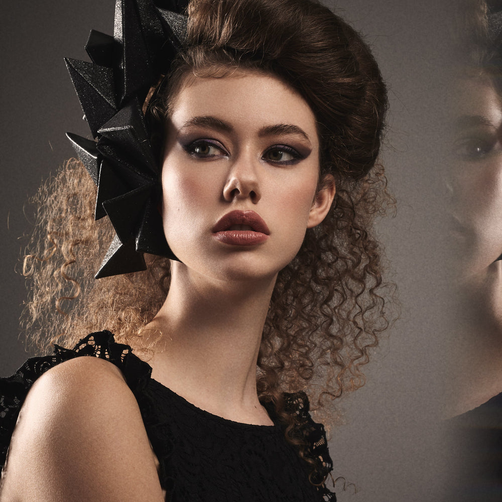 
                  
                    A woman in a black dress wears a black geometric headpiece on the left side of her head. 
                  
                