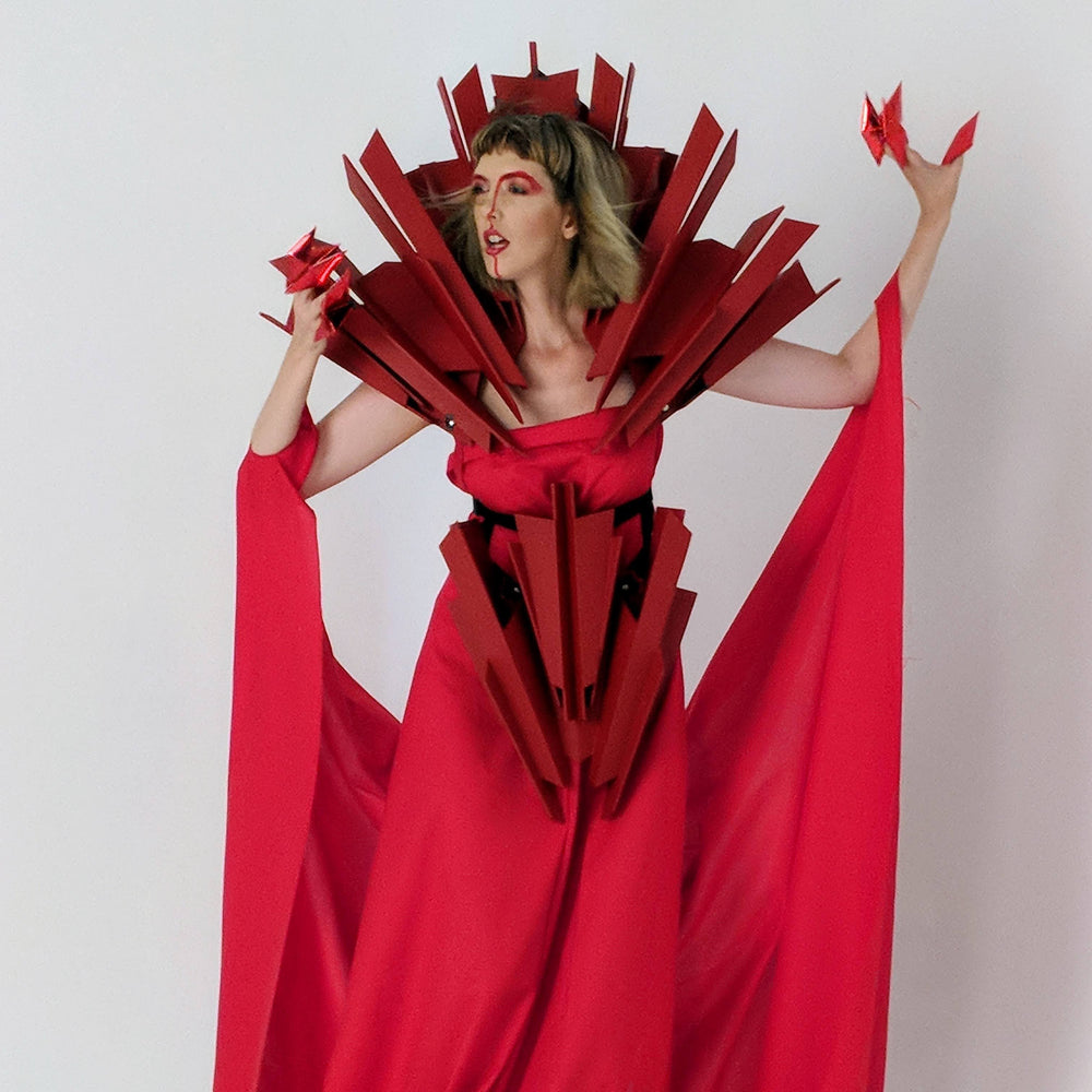 A model poses in an avant-garde red dress with sharp, angular accents, creating a dramatic, futuristic silhouette.