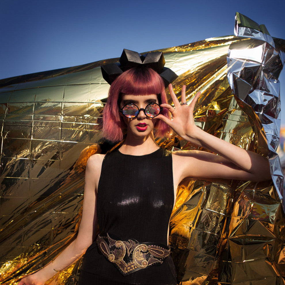 
                  
                    A model with pink hair wears a black sleeveless dress, black crown, and an ornate belt, posing against a shiny gold backdrop.
                  
                