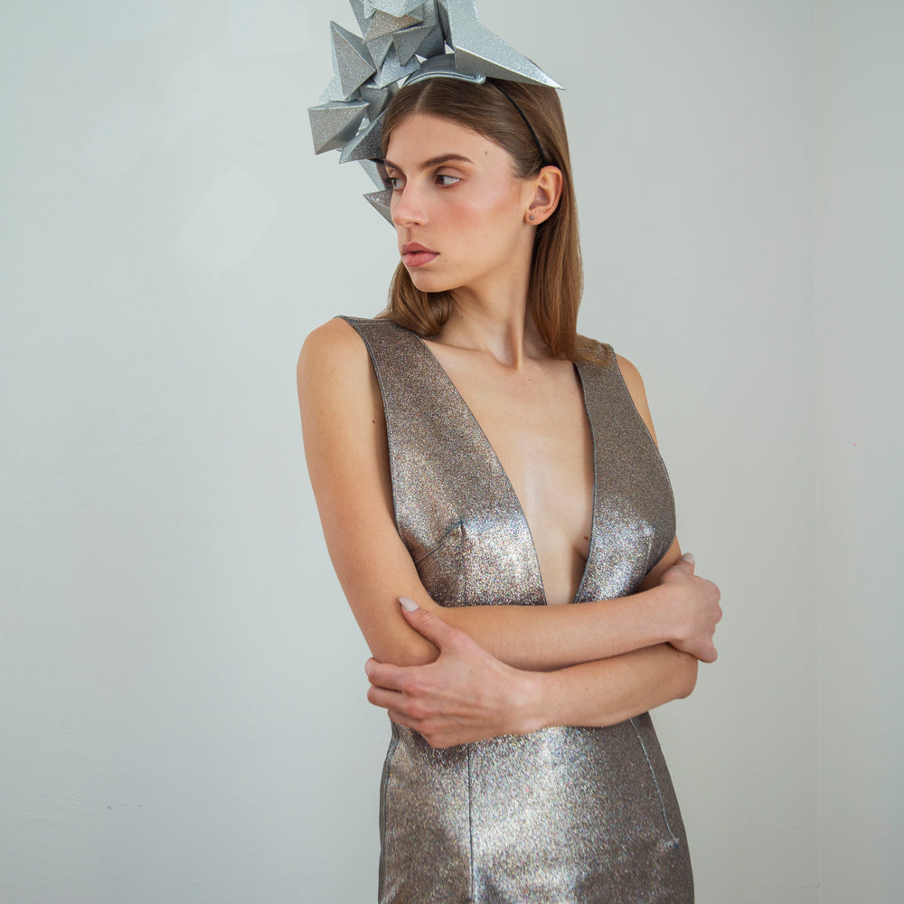 A woman in a silver dress stands in front of a white background wearing a large silver geometric headpiece. 