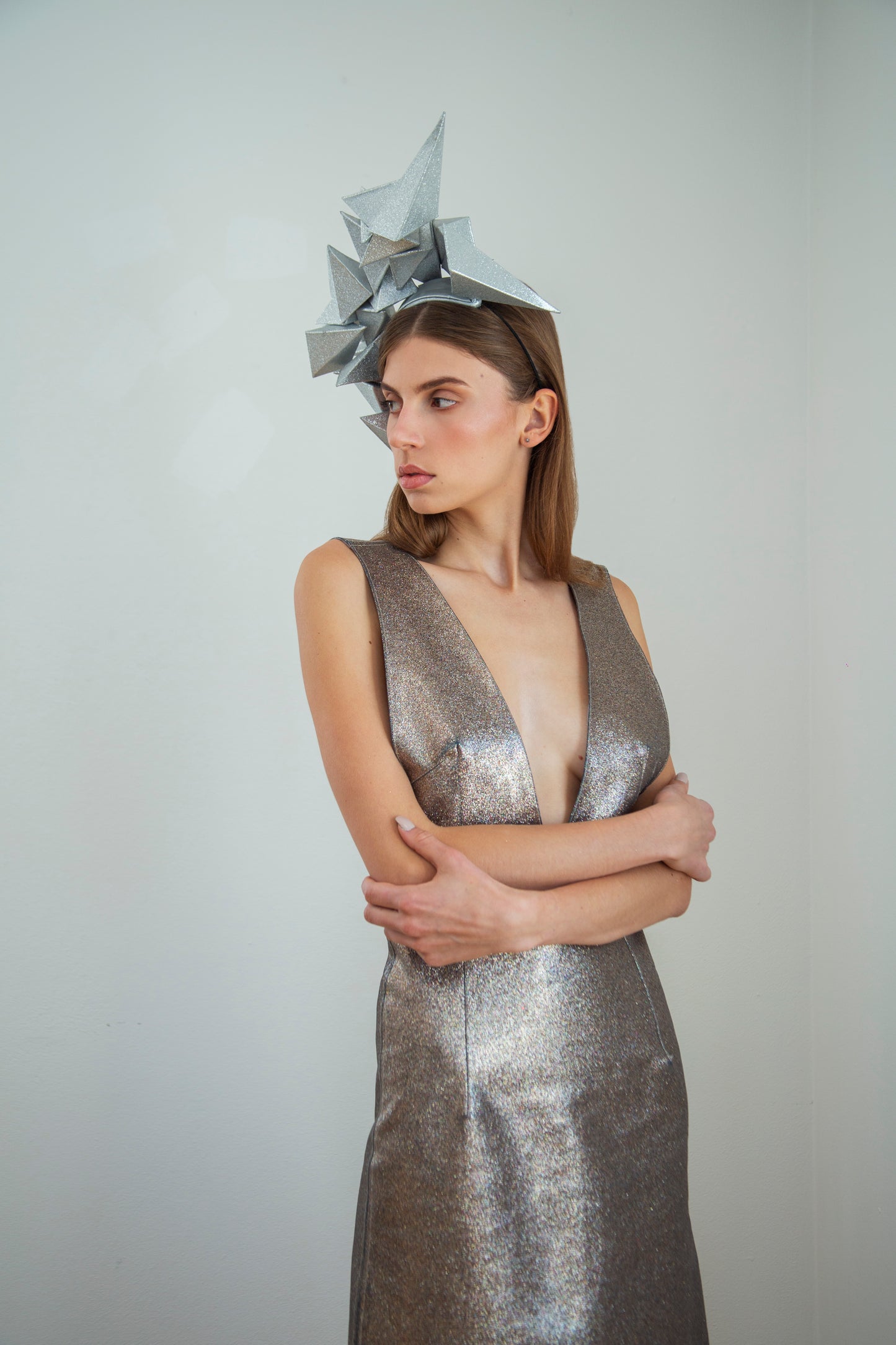 A woman in a silver dress stands in front of a white background wearing a large silver geometric headpiece. 