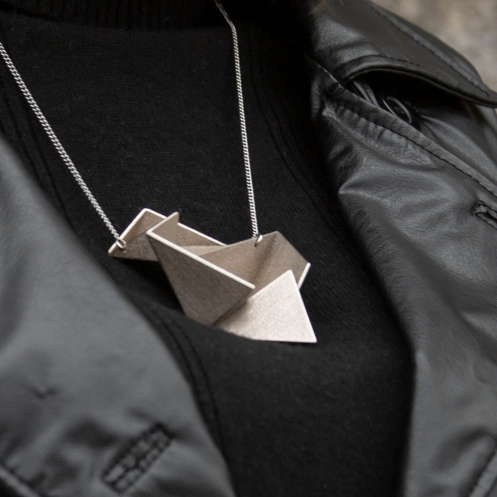 
                  
                    A geometric, origami-inspired pendant in metallic beige hangs from a silver chain, worn over a black turtleneck and leather jacket.
                  
                