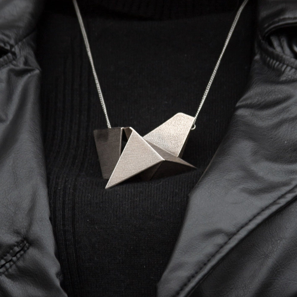 A geometric, origami-inspired pendant in metallic beige hangs from a silver chain, worn over a black turtleneck and leather jacket.