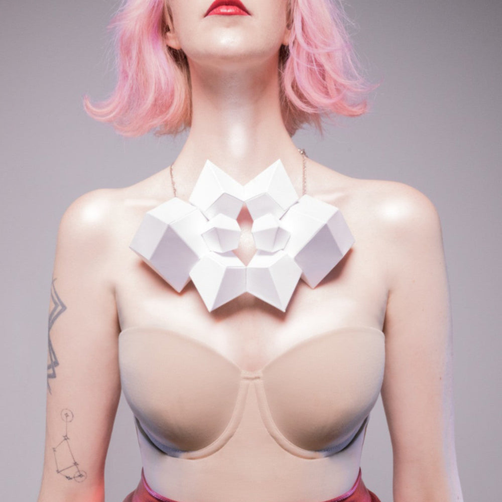 A model with pink hair showcases a geometric white necklace over a beige top, enhancing her bold red lips and tattooed arm.