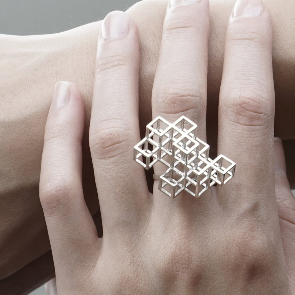 
                  
                    A close-up of a silver geometric ring made of interlocking cubes worn on a hand, showcasing intricate 3D design and a polished finish.
                  
                