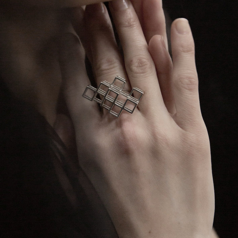 
                  
                    A close-up of a hand wearing a modern, geometric silver ring with interwoven square shapes, highlighting elegant design and craftsmanship.
                  
                