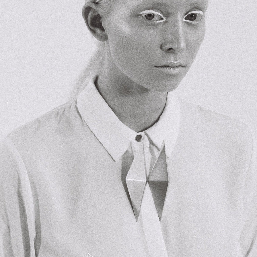 A person wears a crisp white shirt with a geometric silver brooch, set against a simple gray background.