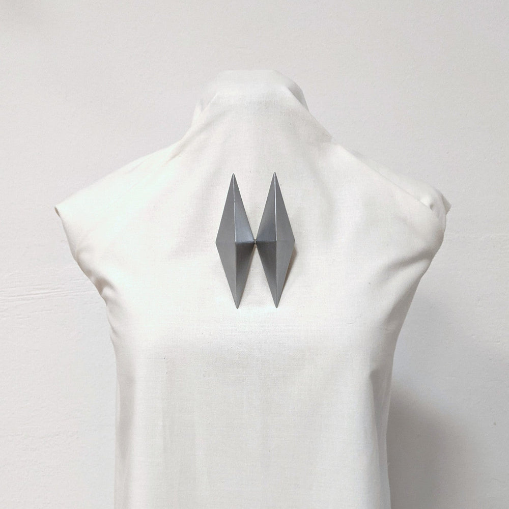 
                  
                    A metallic geometric sculpture resembling a bowtie, mounted on the back of a white mannequin torso against a plain background.
                  
                