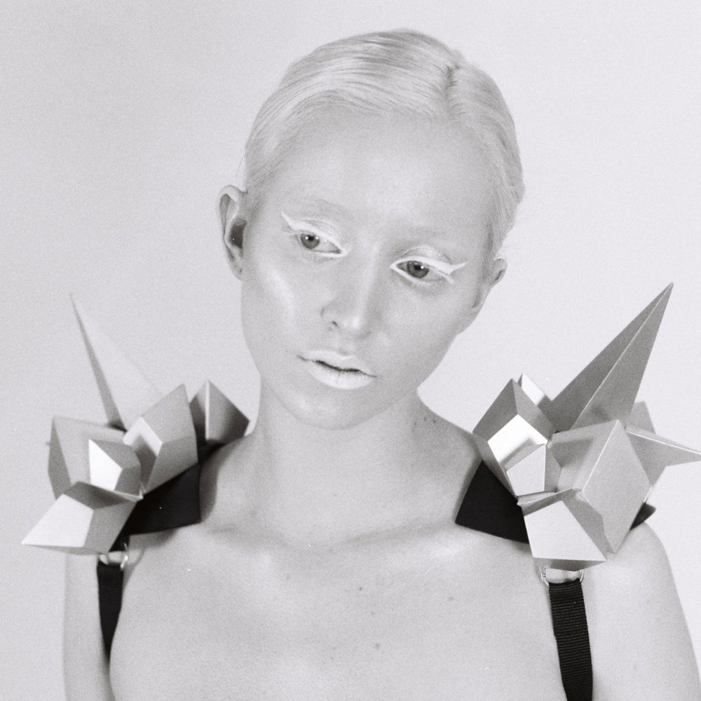 A model wears a striking, geometric shoulder piece with sharp, angular shapes, showcasing innovative fashion design in black and white.
