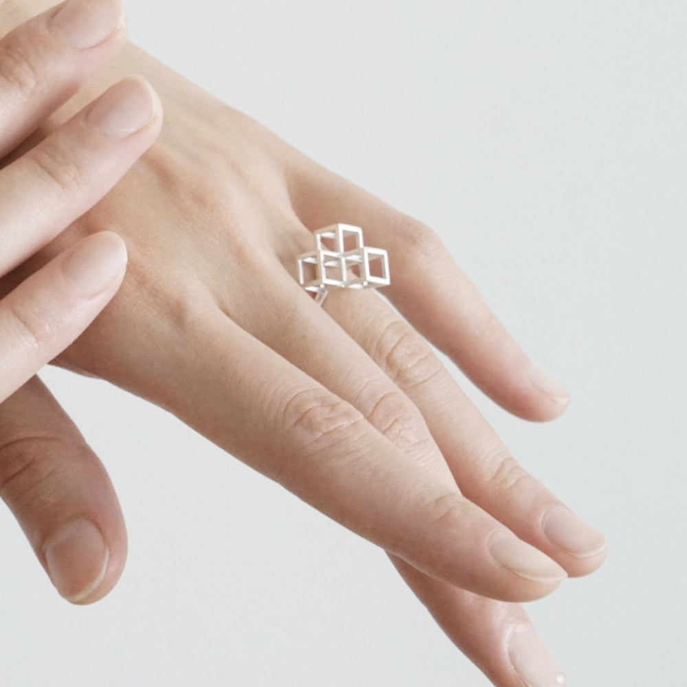 A close-up of a hand wearing a modern silver ring with geometric cube designs against a light background.