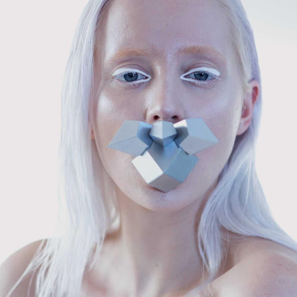 A close-up image of a person with long, straight white hair against a light background, displaying bare shoulders and holding a geometric shape in her mouth. 