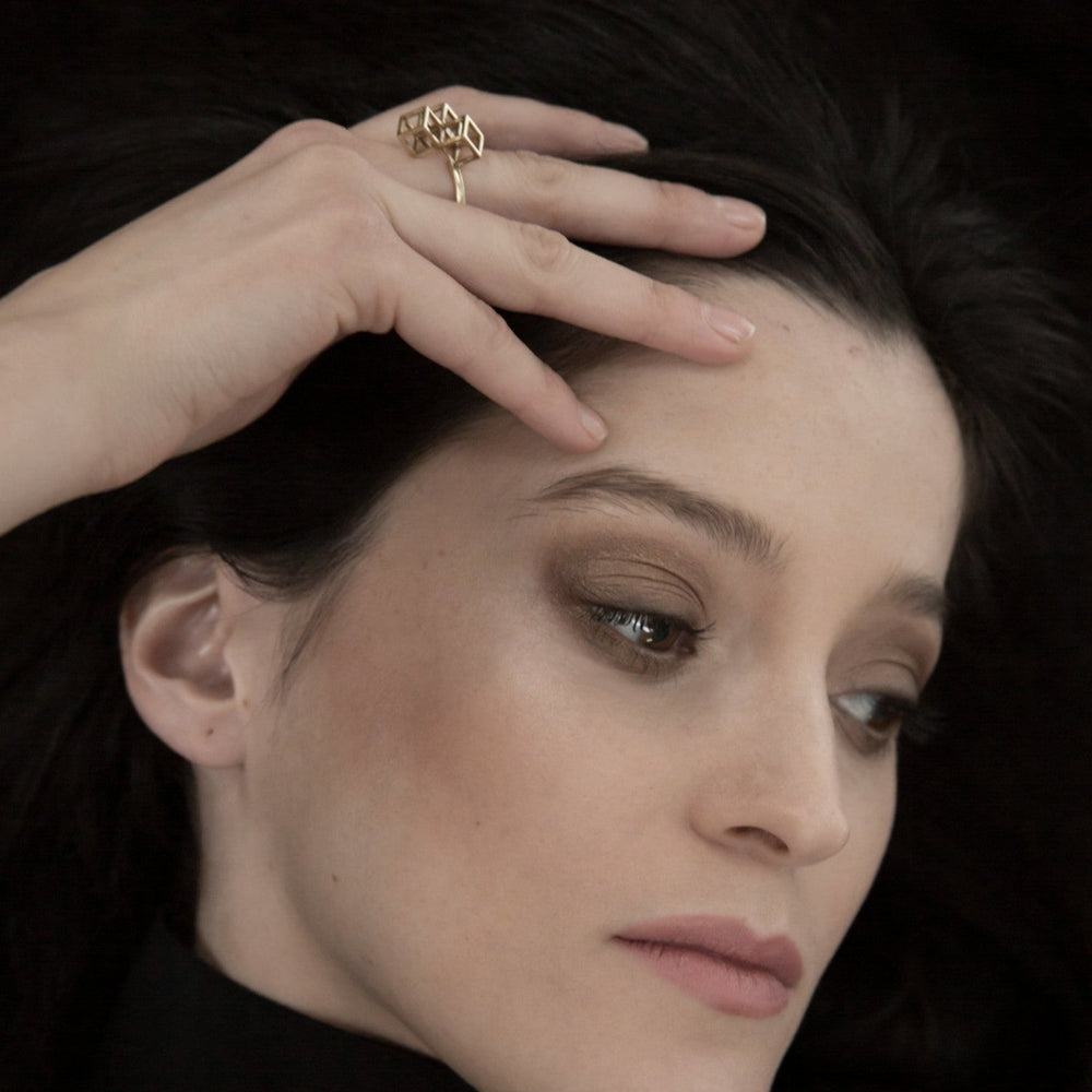 
                  
                    A close-up of a hand gracefully touching dark hair, showcasing a stylish geometric gold ring on the finger against a muted background.
                  
                