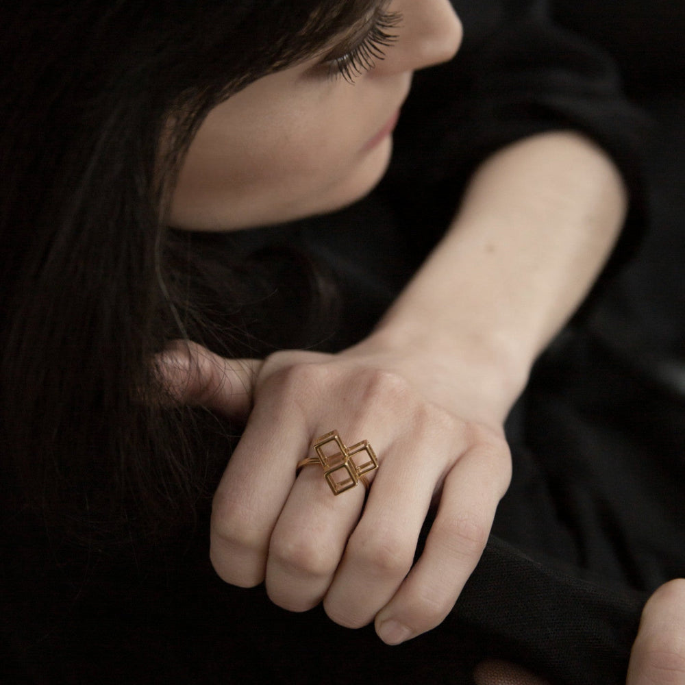 
                  
                    A close-up of a hand wearing a geometric gold ring, resting casually against a dark garment.
                  
                