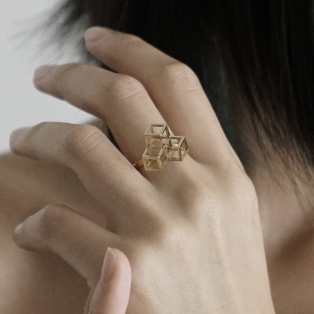 A close-up of a hand wearing a geometric gold ring featuring overlapping square shapes, with a soft focus on the background.