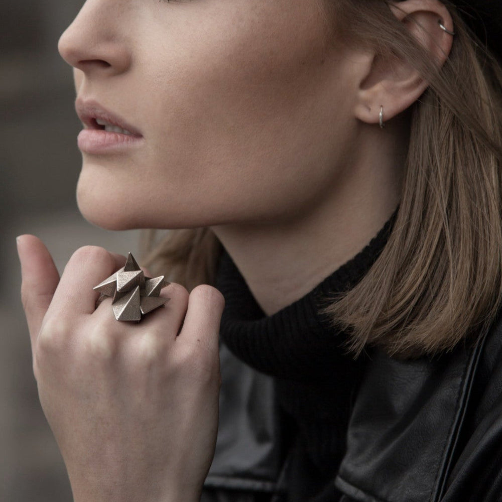 
                  
                    A close-up of a woman's hand showcasing a unique, geometric ring made of metal, against a backdrop of urban settings.
                  
                