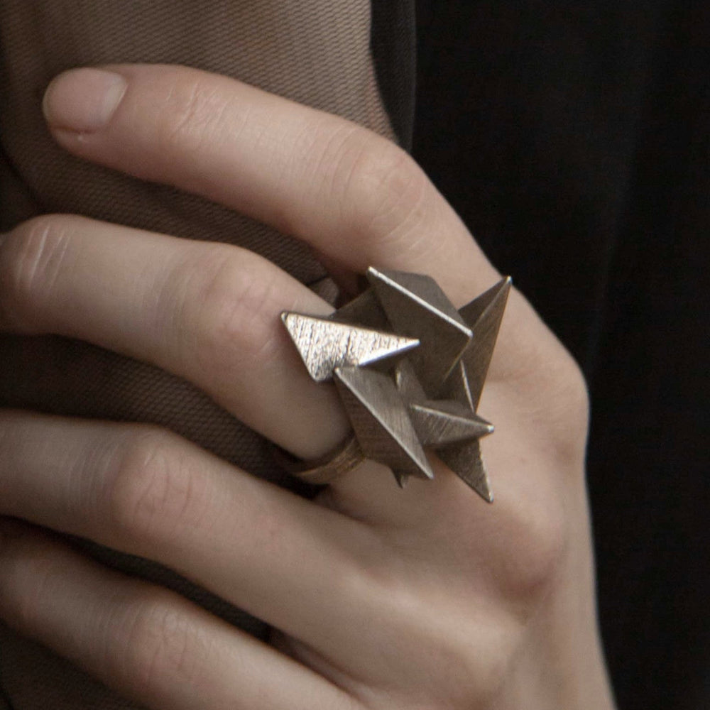 A close-up of a woman's hand showcasing a unique, geometric ring made of metal, against a black background.