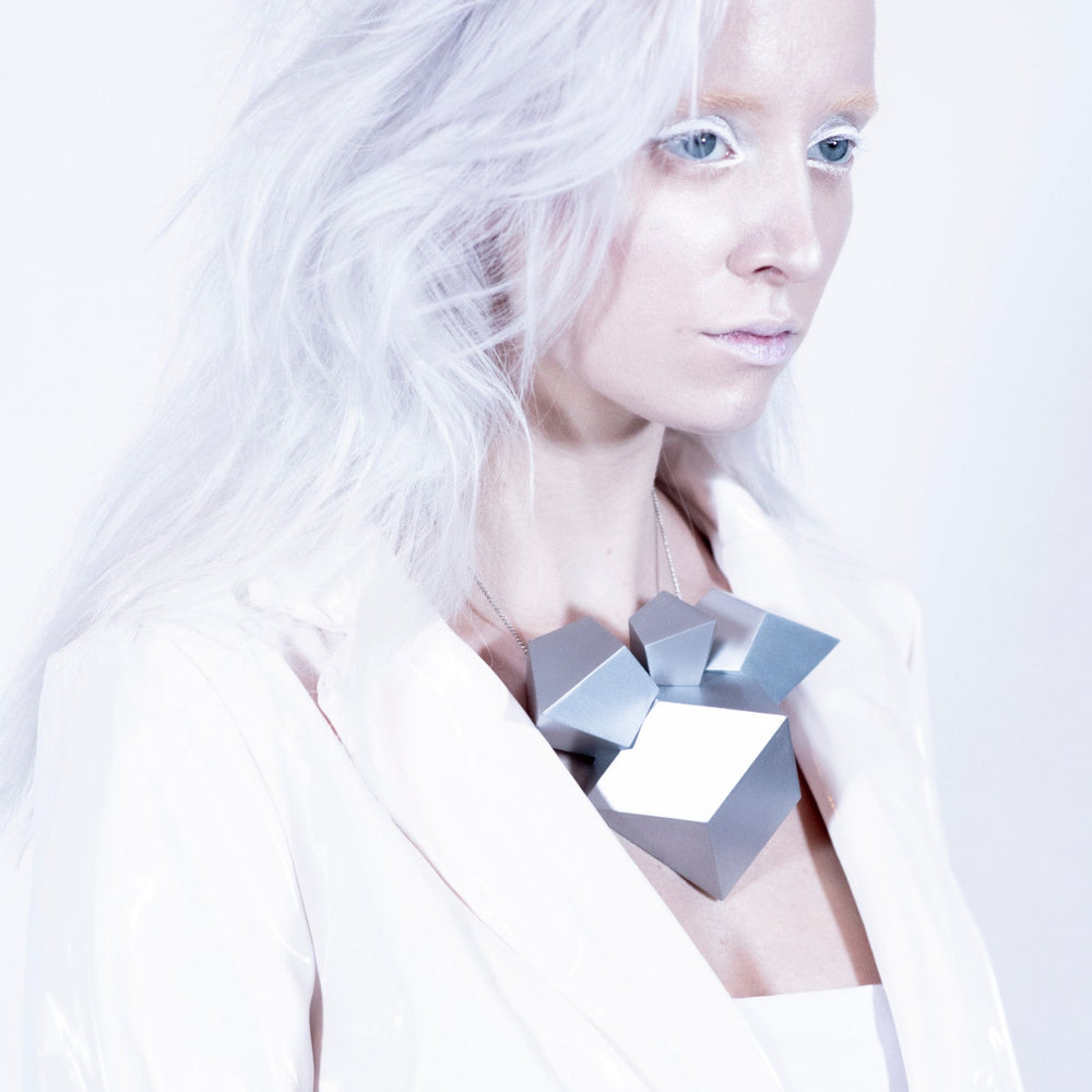 A model with long, wavy platinum hair wears a striking geometric silver necklace and a glossy white blazer against a light background.