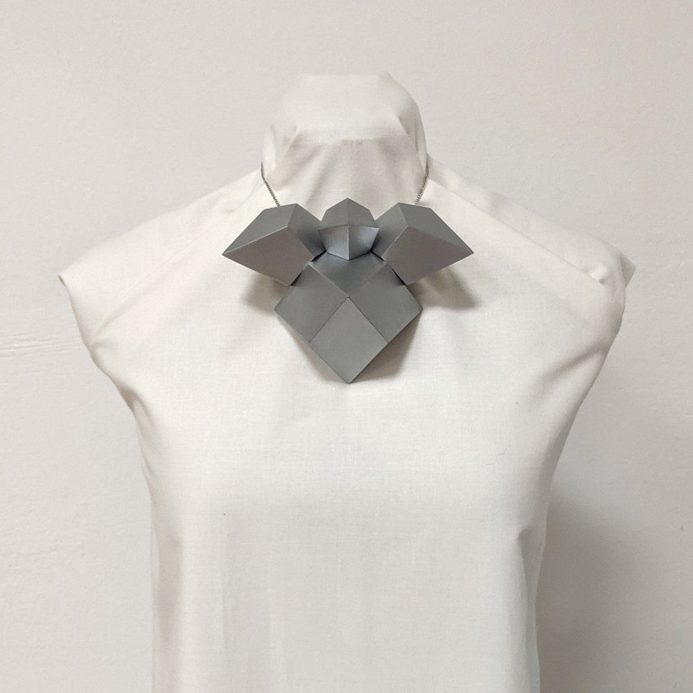 
                  
                    A sculptural, geometric silver necklace displayed on a dress form, featuring angular shapes and a modern design.
                  
                