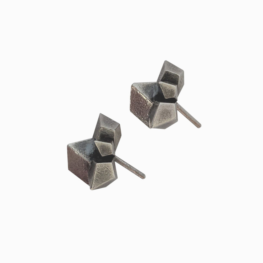 
                  
                    A pair of geometric silver earrings with textured surfaces and angular shapes, resting on a white background.
                  
                