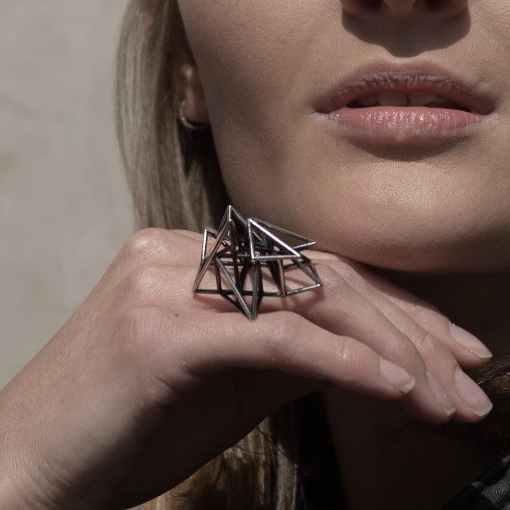 A close-up of a woman's hand holding a geometric metal ring, showcasing its sharp angles against her soft facial features.
