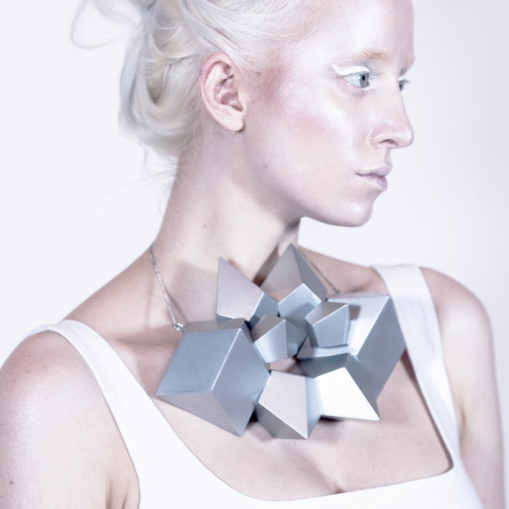 
                  
                    A close-up of a person's neck adorned with a striking, geometric silver necklace featuring various angular shapes.
                  
                
