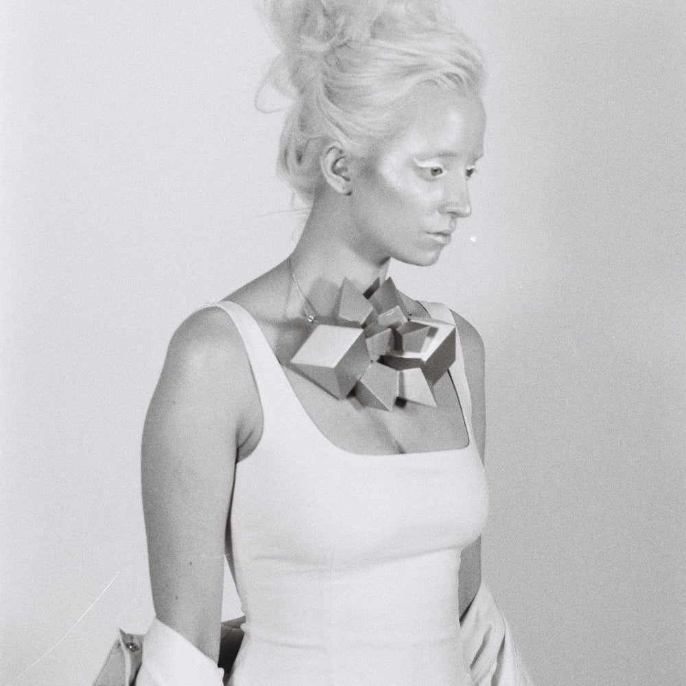 
                  
                    A person in a fitted white dress adorned with a geometric statement necklace, styled with elegant, tousled blonde hair.
                  
                