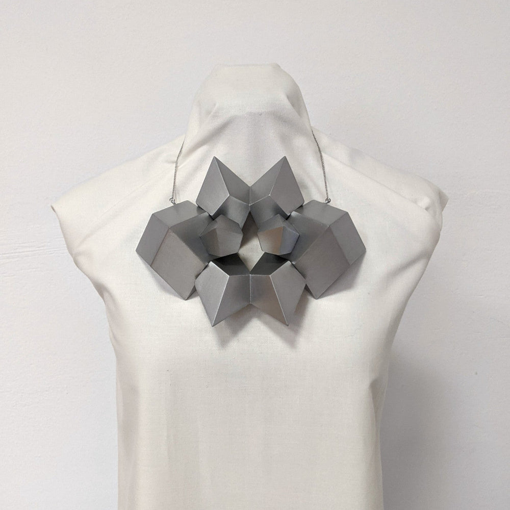 
                  
                    A sculptural silver necklace featuring geometric shapes arranged in an intricate, star-like design on a mannequin bust.
                  
                