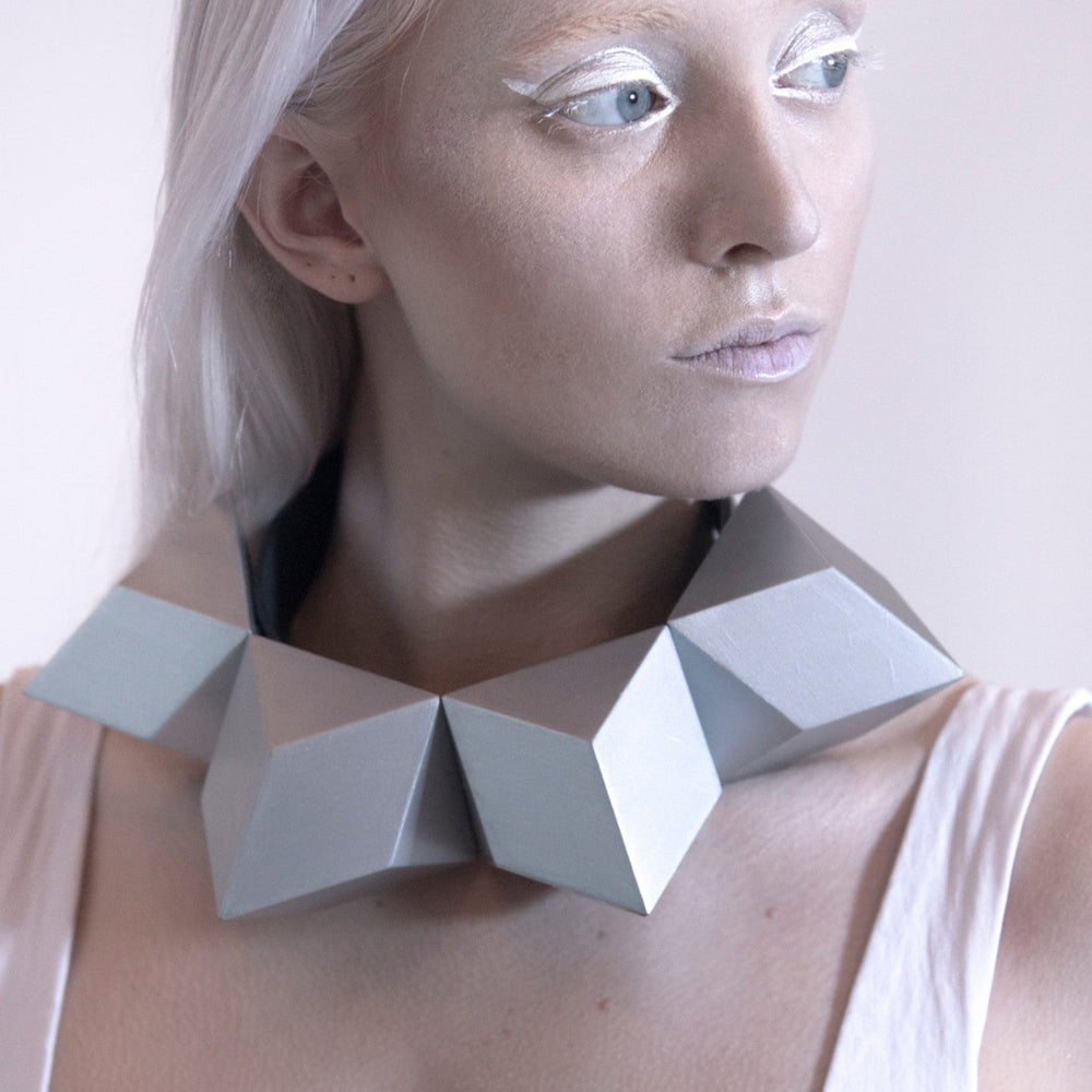 A person wears a bold, geometric silver necklace made of angular, three-dimensional shapes, contrasting with a simple white top.