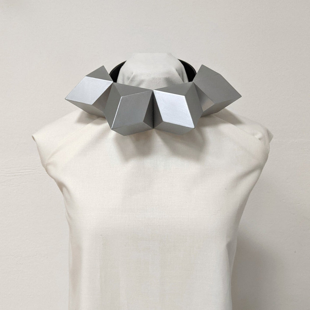 
                  
                    A sculptural silver geometric necklace displayed on a dress form, featuring multiple angular shapes and a sleek black band.
                  
                