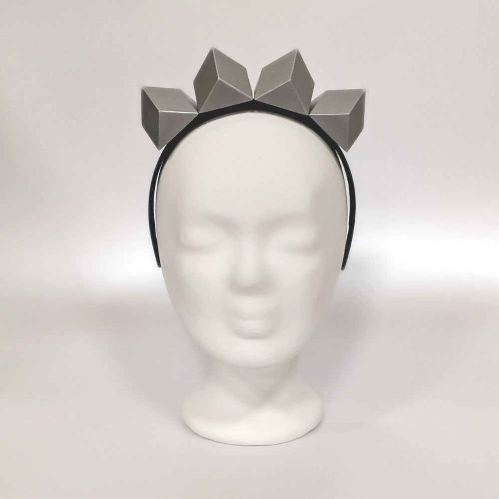 A headband featuring a group of geometric, grey shapes resembling a crown or tiara, poised atop a plain, white display head.