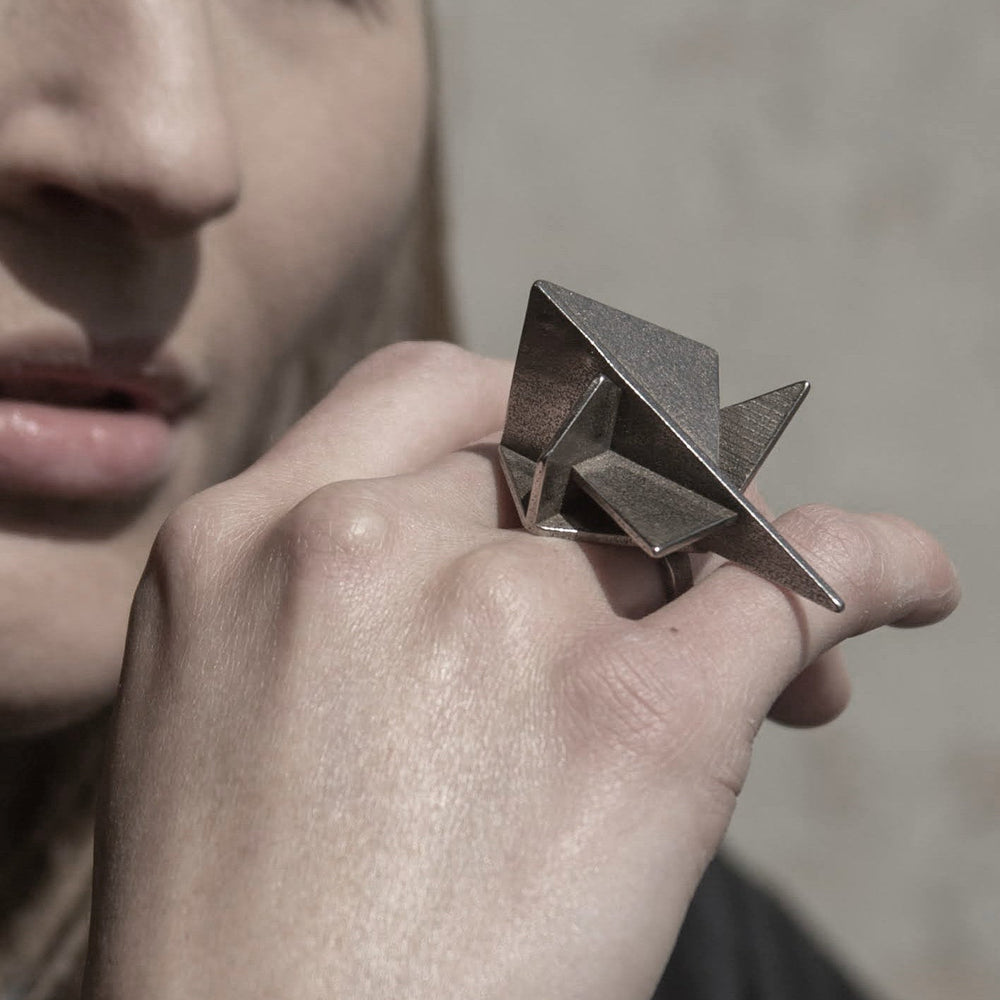 
                  
                    A person wearing a metallic geometric ring, showcasing angular shapes and a polished finish against a blurred background.
                  
                