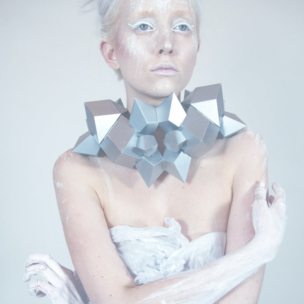 A figure with silver geometric jewelry around the neck, adorned with white body paint, set against a soft, pale background.
