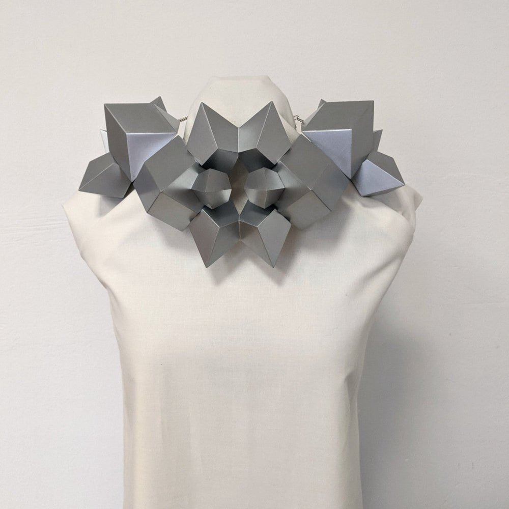 A geometric silver necklace made of angular shapes displayed on a mannequin against a plain background.