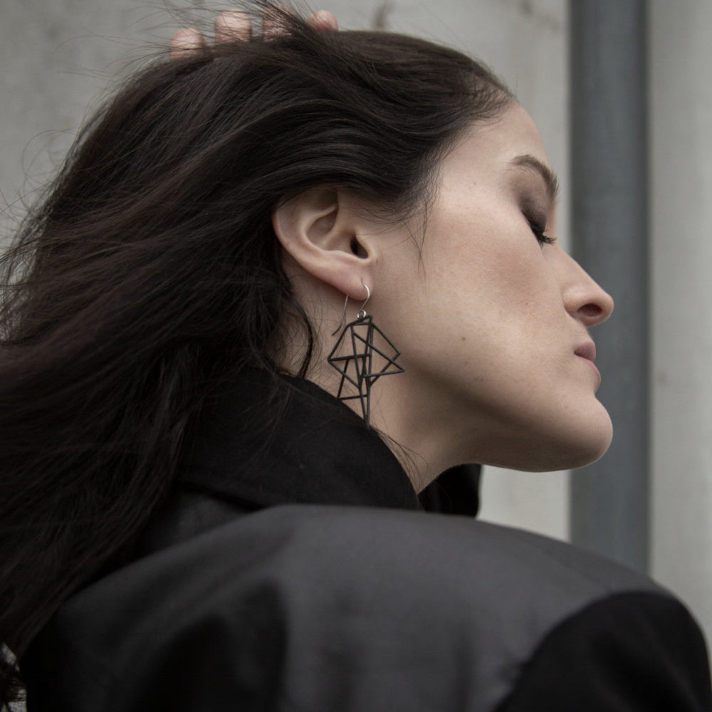 A woman with long, dark hair wears stylish geometric earrings and a black jacket, posing against a textured, neutral background.