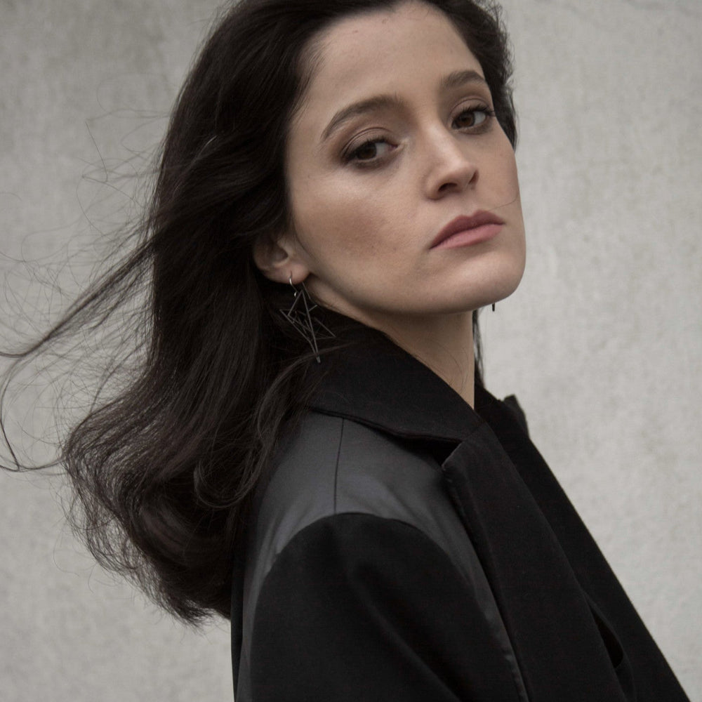 
                  
                    A woman with long dark hair wears bold geometric earrings, styled in a chic black jacket, against a minimalist background.
                  
                