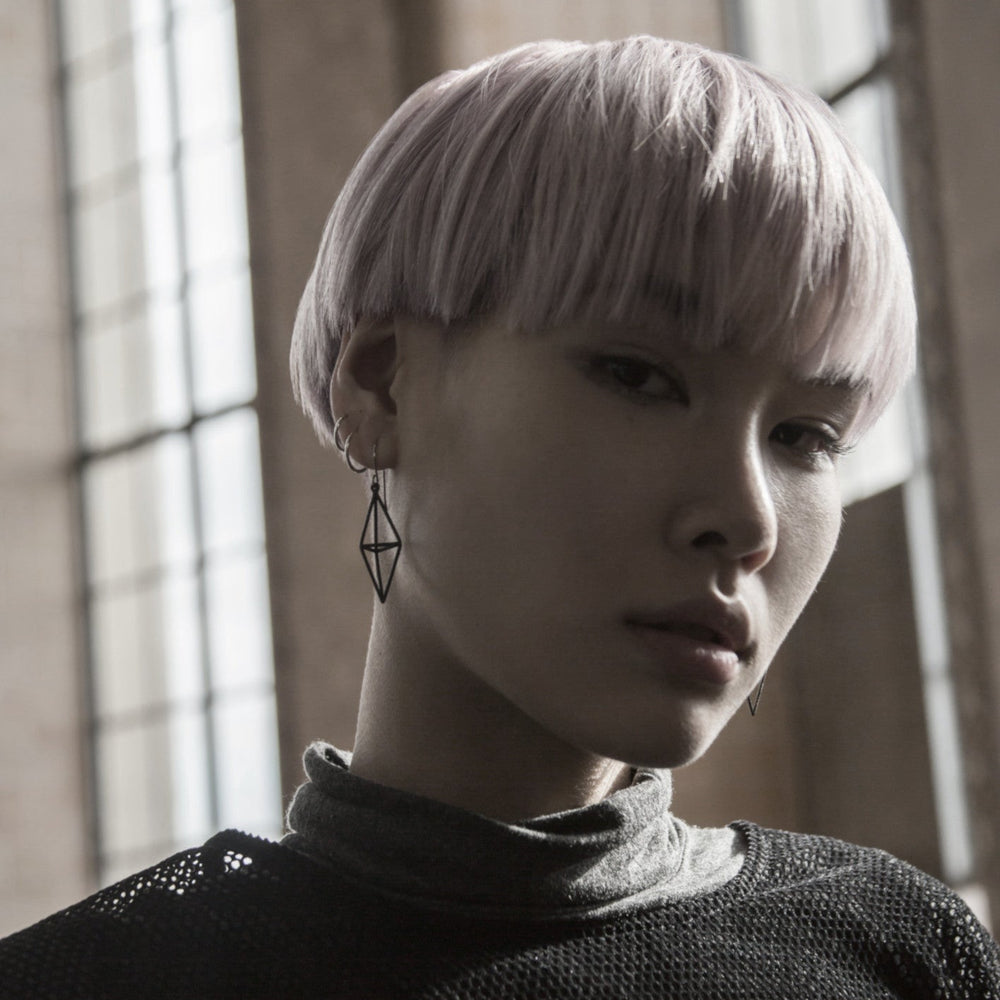 
                  
                    A person with short, pastel pink hair wears a geometric earring and a gray turtleneck top, captured in soft natural lighting.
                  
                