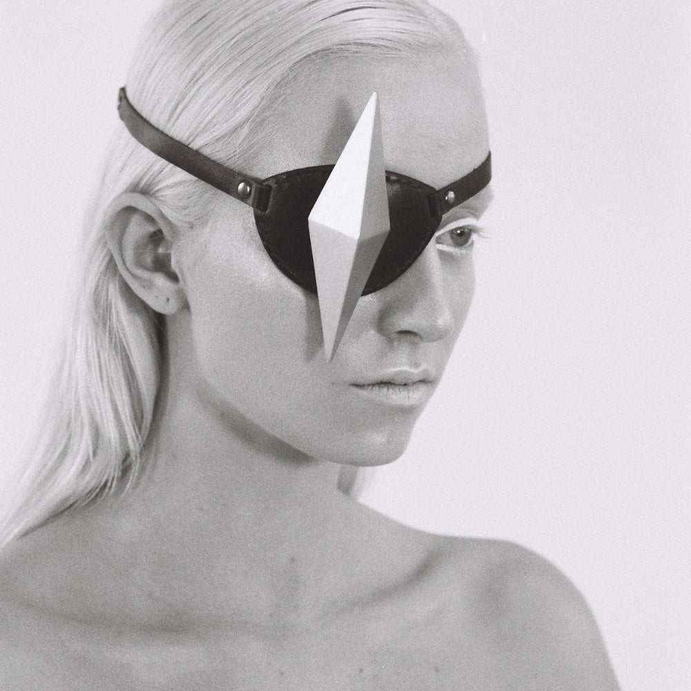 A close-up of a person with long, straight, platinum blond hair, wearing a black eyepatch with a silver geometric shape and showcasing a bare shoulder in black and white.