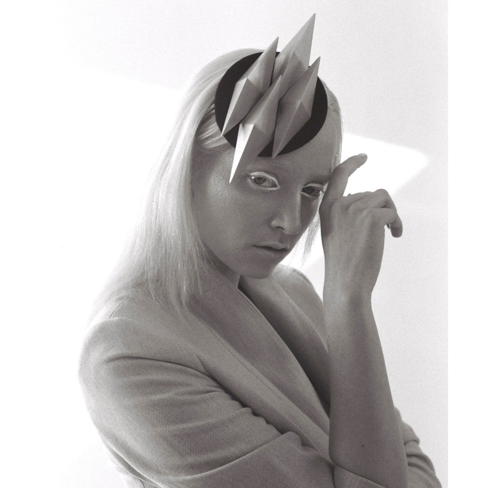 A figure in a grey blazer poses with one hand raised, showcasing an artistic headpiece of angular shapes against a minimalist backdrop.