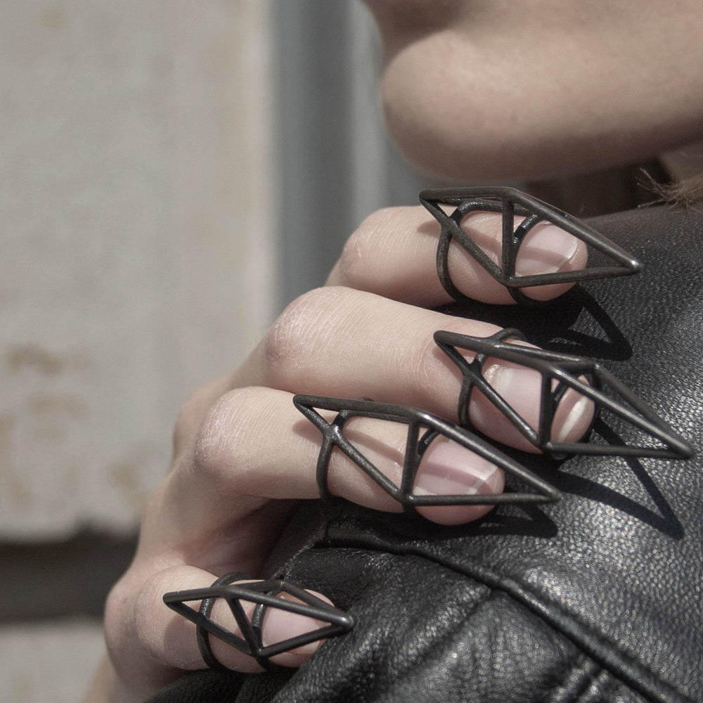 A close-up of a hand with black geometric rings, resting on a leather jacket, showcasing artistic nail design and a stylish pose.