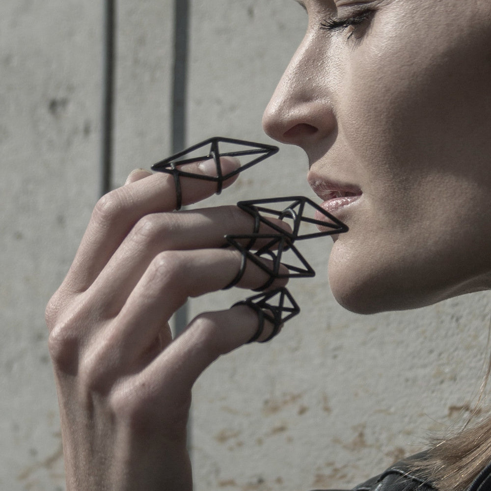 
                  
                    A delicate hand adorned with geometric black wire rings, set against a textured concrete wall, showcasing a trend-forward accessory style.
                  
                
