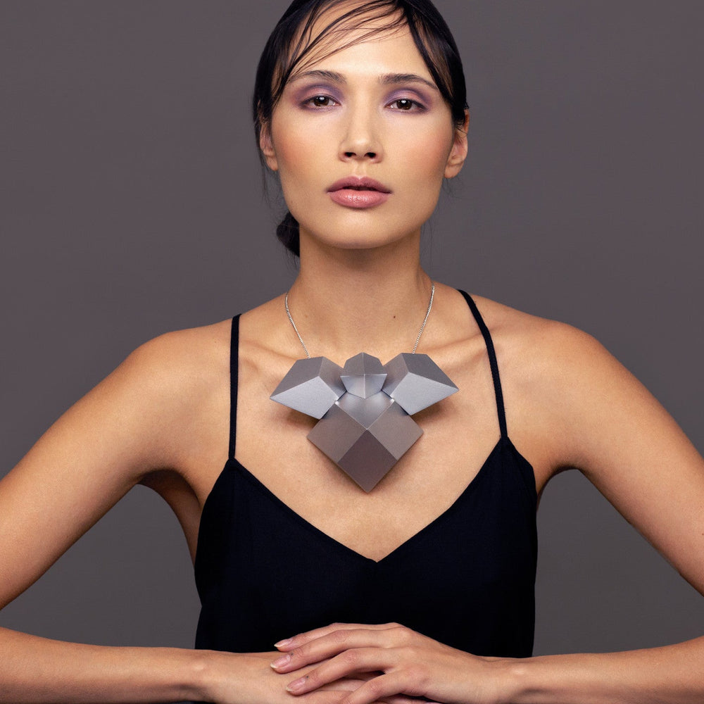 
                  
                    A model wears a bold geometric necklace made of silver and grey shapes, paired with a sleek black top against a neutral background.
                  
                