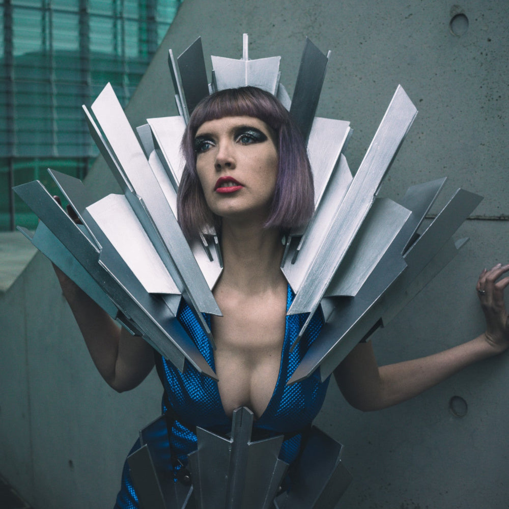 A figure in a futuristic blue outfit is adorned with sharp metallic protrusions, set against a modern architectural backdrop.