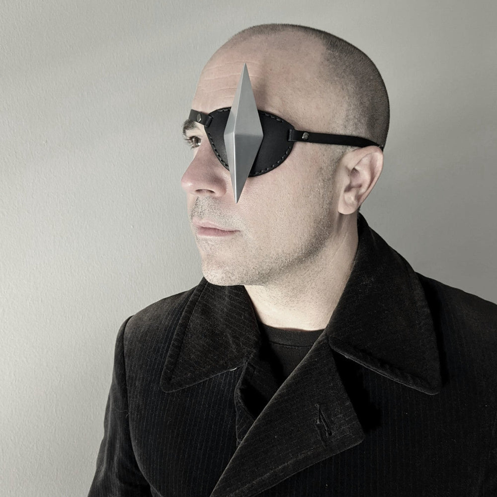 A person with a bald head, wearing a dark corduroy coat and black eyepatch with a silver geometric shape stands against a light gray wall, creating a minimalist look.