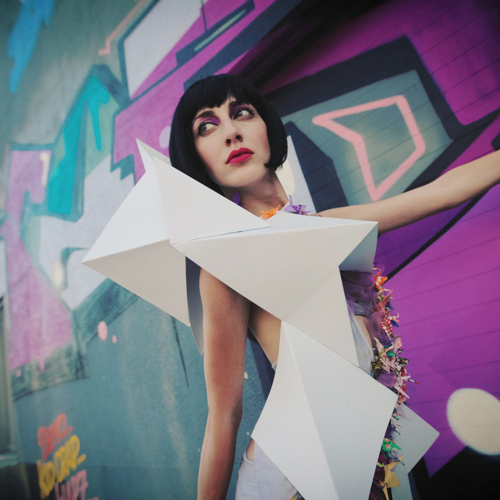 A model showcases an avant-garde outfit featuring geometric white shapes and colorful embellishments against a vibrant graffiti backdrop.