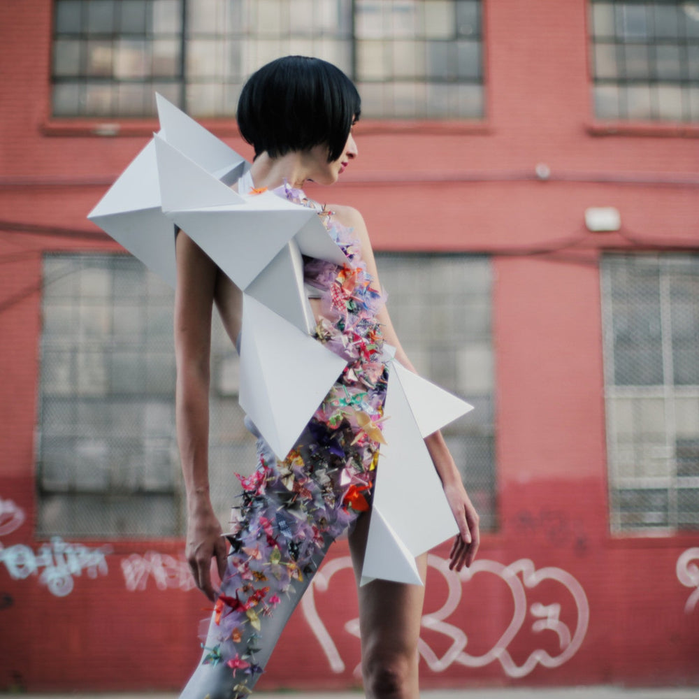 A model poses in an artistic outfit adorned with colorful paper elements and geometric white shapes against a red brick background.