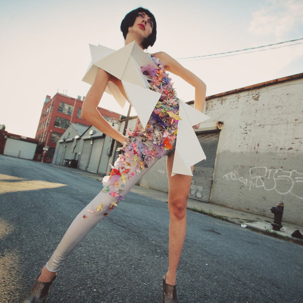 
                  
                    A model poses in an urban setting, showcasing a unique outfit with origami-like shapes and colorful floral embellishments.
                  
                