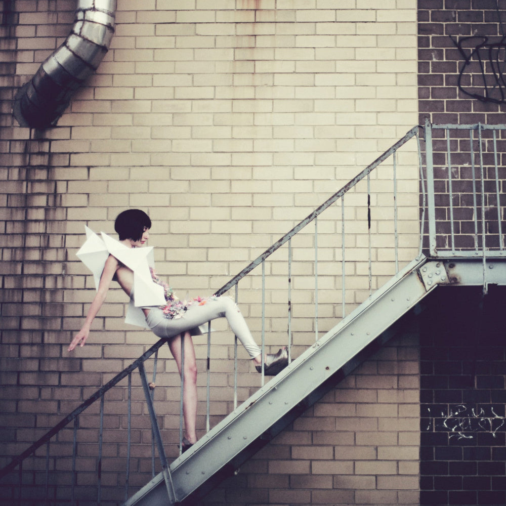 
                  
                    A figure in a silver bodysuit and geometric body sculpture descends a metal staircase against a brick wall.
                  
                