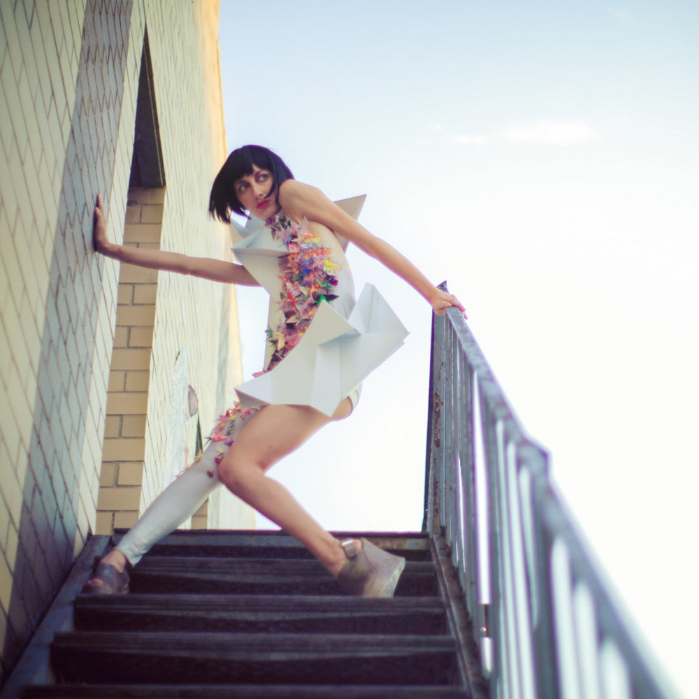 
                  
                    A person in a colorful, avant-garde outfit climbs a staircase against a backdrop of a sunlit sky and brick wall.
                  
                