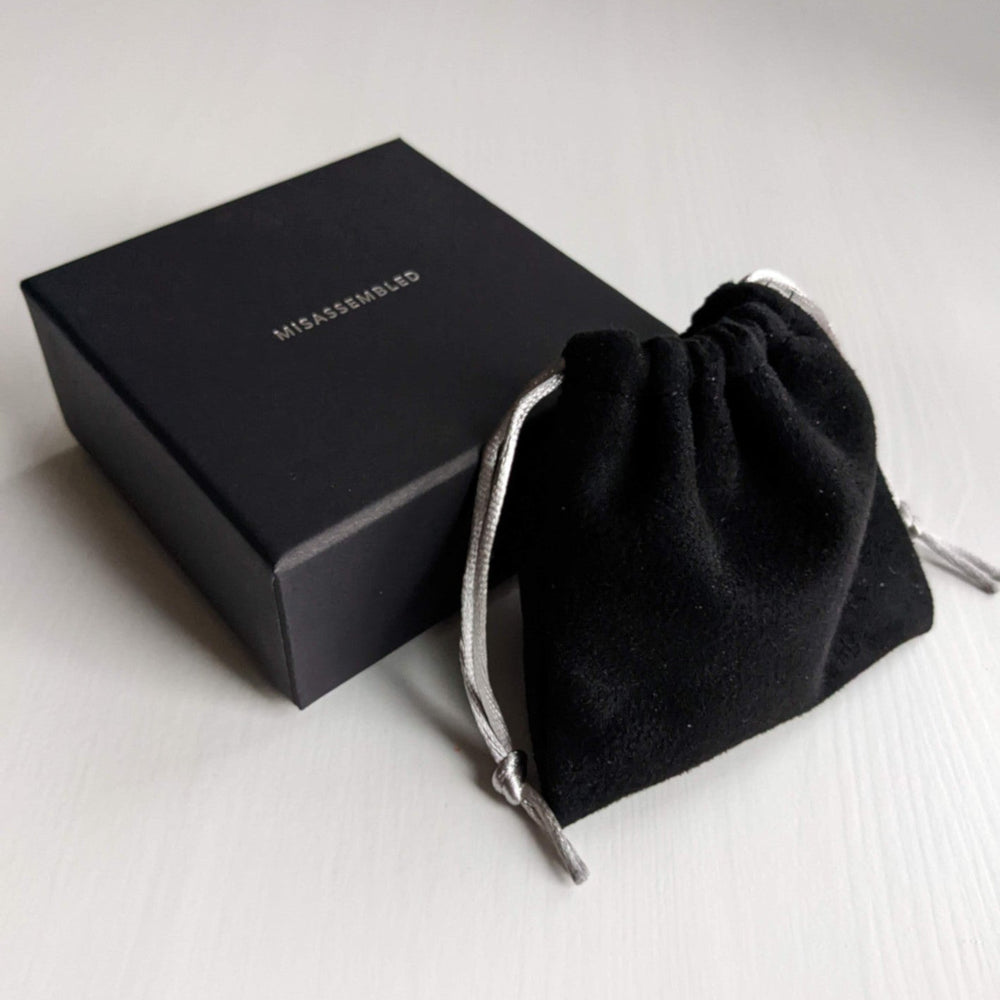 
                  
                    A sleek black gift box with "MISASSEMBLED" printed on top, next to a small black velvet pouch with silver drawstrings.
                  
                